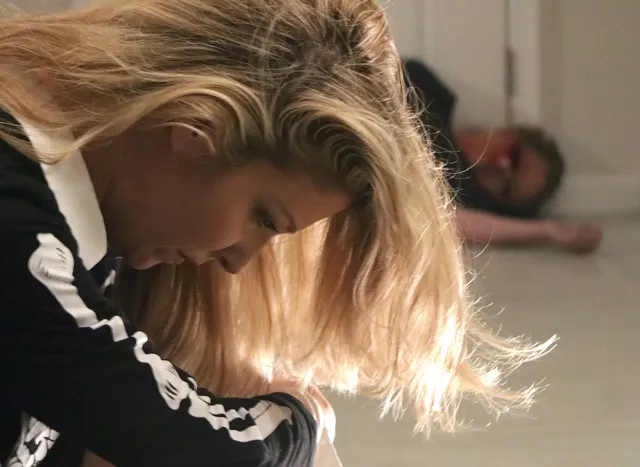 Actress with blonde hair looks down intensely during a dramatic scene filmed inside a house, with another actor lying on the floor in the background