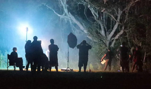 Silhouettes of a film crew working at night on an outdoor set with a bright light on a stand, haze, and Spanish moss hanging from trees