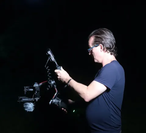 Cinematographer operates a camera mounted on a gimbal stabilizer with an attached shotgun microphone during a nighttime outdoor shoot