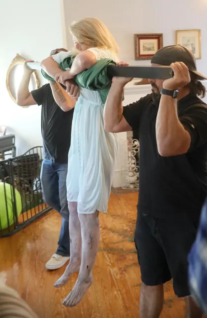 Crew members carry an actress in a nightgown past the camera while rigging equipment inside a house during a film production