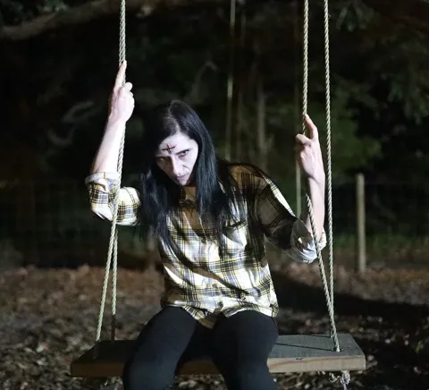 Actress in horror makeup with a bloody cross on her forehead and pale skin sits on a rope swing at night during filming