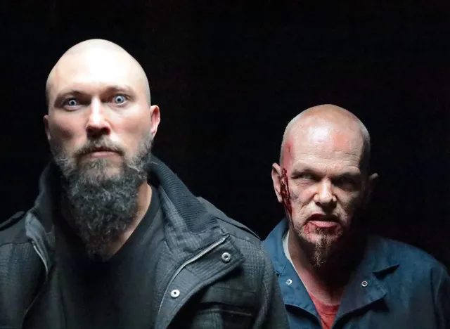 Two actors in character on a dark set, one with a beard in a black jacket and another with blood effects makeup on his face