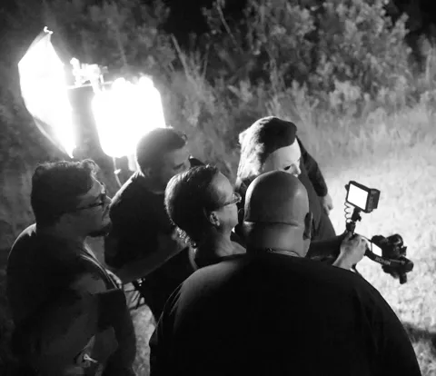 Black and white shot of the film crew huddled together reviewing footage on a camera monitor during a nighttime outdoor shoot with a large softbox light