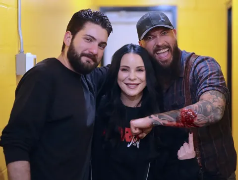 Three smiling cast and crew members pose together between takes, one showing off a prosthetic bloody wound on his arm