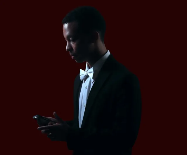 Dramatic low-key portrait of a young man in a black tuxedo and white bow tie, lit from the side against a dark red background
