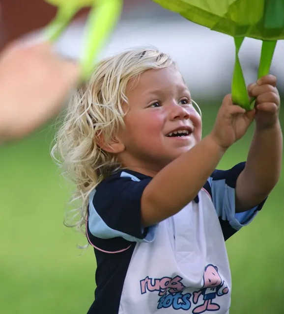 Smiling young child with curly blonde hair in a rugby jersey reaches up to grab a green parachute toy outdoors