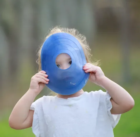 Playful toddler in a white top peeks through the hole of a blue disc toy held up to her face outdoors