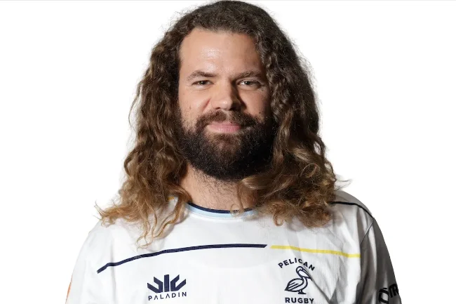 Headshot of a bearded man with long curly hair wearing a white Pelican Rugby jersey against a white studio background