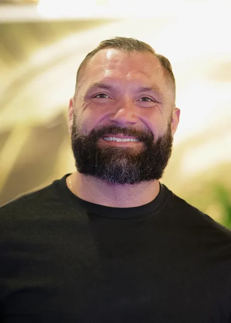 Warm portrait of a smiling bearded man in a black crew neck shirt with soft natural light and tropical foliage in the background