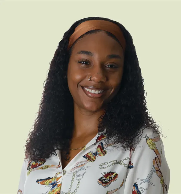 Bright professional headshot of a smiling woman with curly black hair and an orange headband wearing a colorful butterfly-print blouse against a light green background
