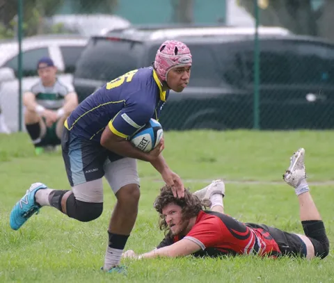 Rugby player in a navy jersey and pink scrum cap breaks away from a diving tackle attempt on a muddy grass pitch