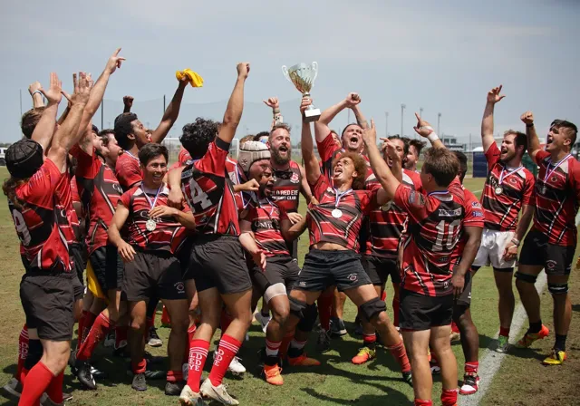 Old Red Eye Rugby Club team celebrates their championship victory, hoisting a trophy and cheering together on the field wearing medals