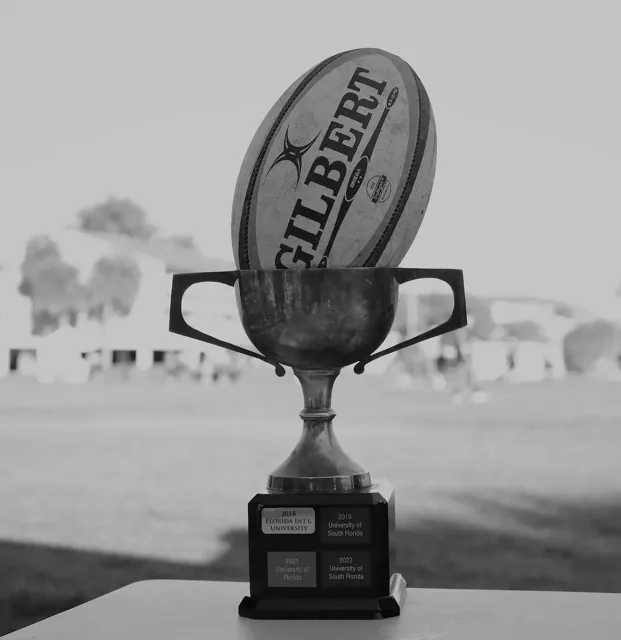 Black and white close-up of a Florida collegiate rugby championship trophy with a Gilbert rugby ball resting on top, engraved with past university winners