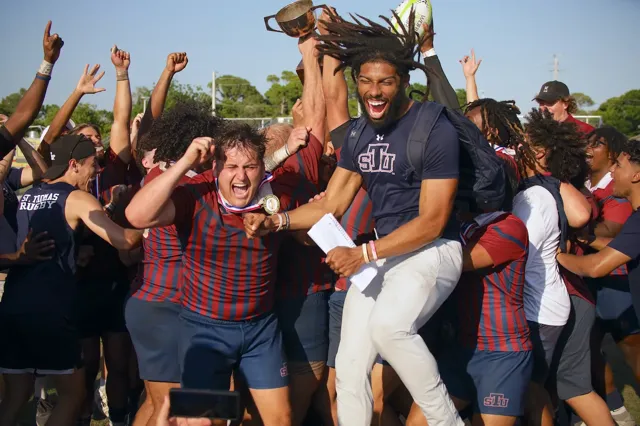 St. Thomas University rugby team erupts in celebration after winning a championship, players jumping and shouting with medals and trophy raised