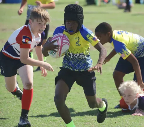 Youth rugby player wearing a protective headguard and Pelican Rugby jersey sprints with the ball past defenders during a tournament match